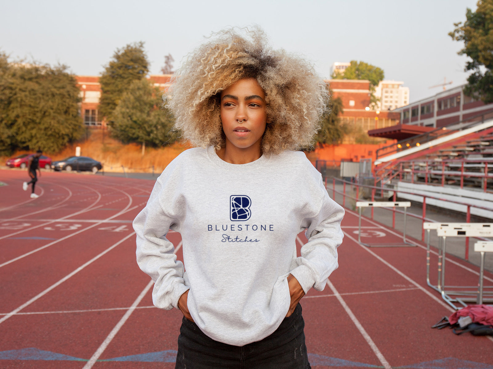 A woman standing on a running track, wearing a grey crewneck with the full Bluestone Stitches logo in blue.